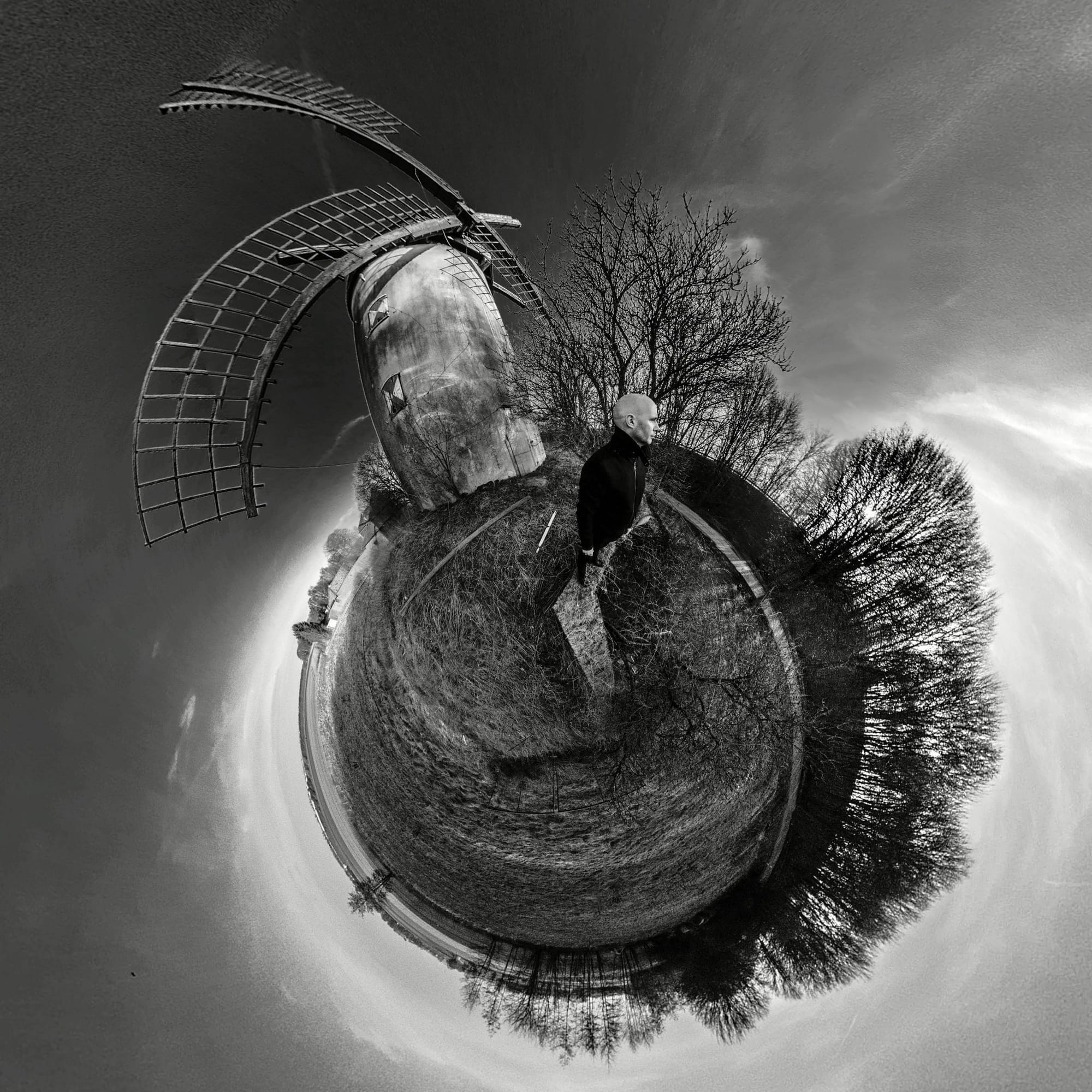 Jens steht in einem verlassenen Dorf, hinter ihm ist eine alte Windmühle zu sehen. Das Bild ist von der Mitte aus aufgenommen und simuliert eine Art Fischaugenoptik.